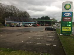A BP forecourt sign promoting Mace and Costa Express, next to a car park, with a building in the distance.