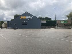 A building, painted grey, with the Subway logo, next to a car park.