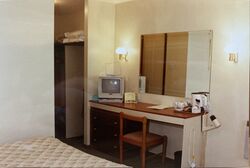 A small hotel room showing a mattress, CRT TV, mirror, desk and chair, wooden drawers and tea and coffee facilities.