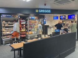 A Greggs store with food chillers, serving counters and tables, positioned within a building.