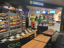A Greggs store with food chillers, serving counters and tables, positioned inside a petrol station.