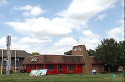 A restaurant building with an overhanging roof, a chimney, red walls, and signs saying Little Chef in a cursive fount.